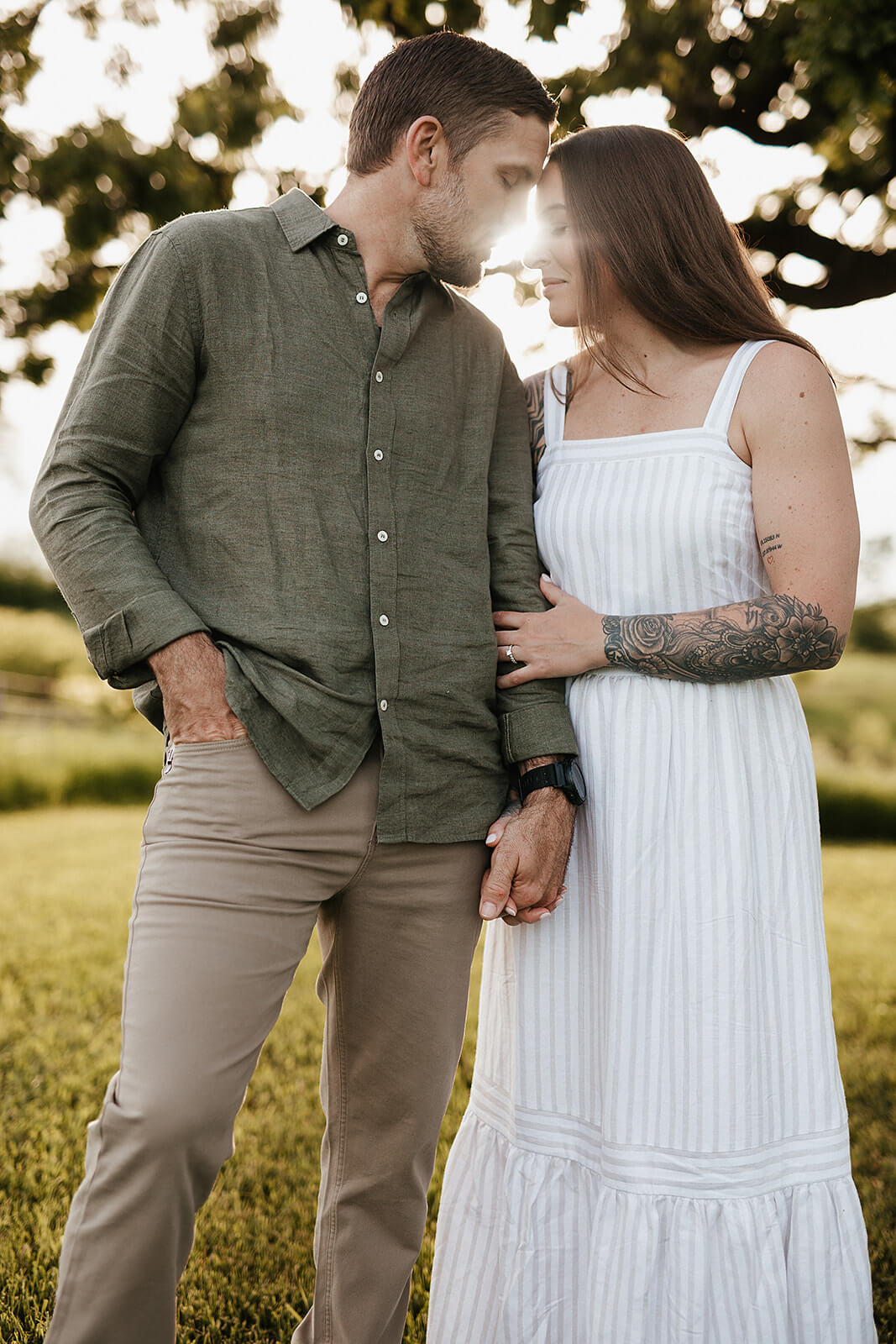 Close-up portrait of a couple standing together outdoors with sunlight filtering through the trees behind them during sunset.