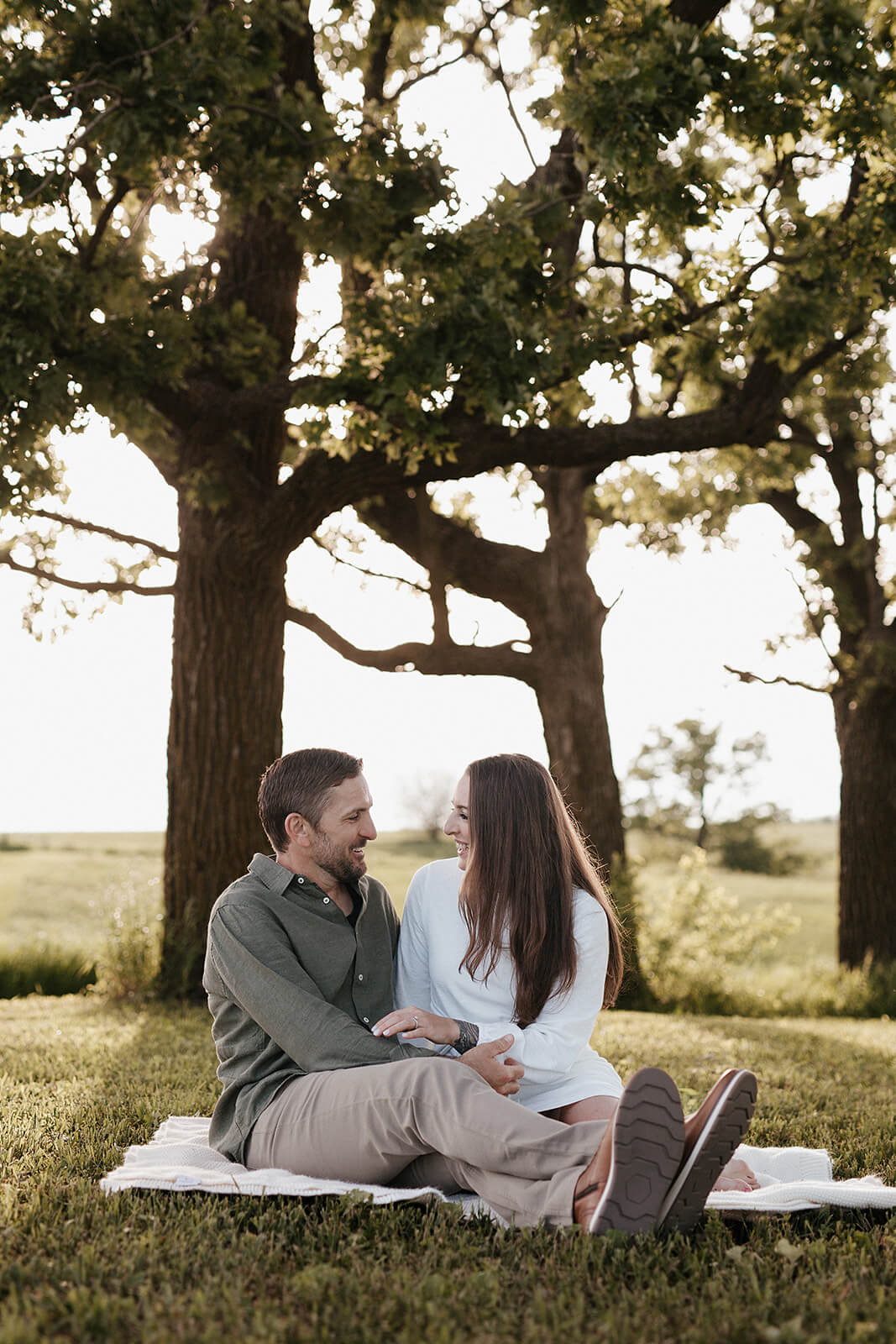 Couple sitting together on a blanket beneath large trees in an open field during sunset with soft golden light shining through the branches.