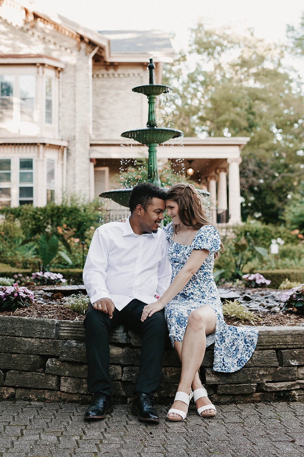 Couple sitting together on a stone wall in front of a fountain and historic home during an outdoor engagement session with flowers and greenery surrounding them.