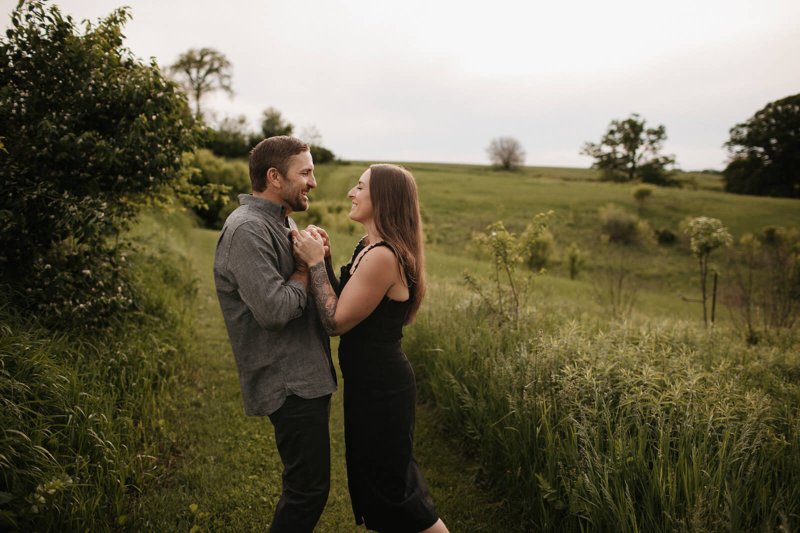 Couple standing together in a grassy field with rolling hills and trees in the background during golden hour, smiling while holding hands.