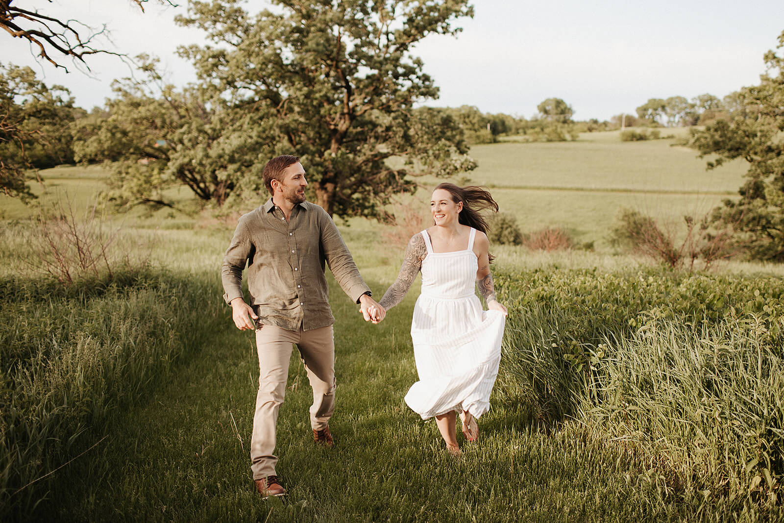 Couple running hand in hand through a grassy field with rolling hills and large trees in the background during golden hour.