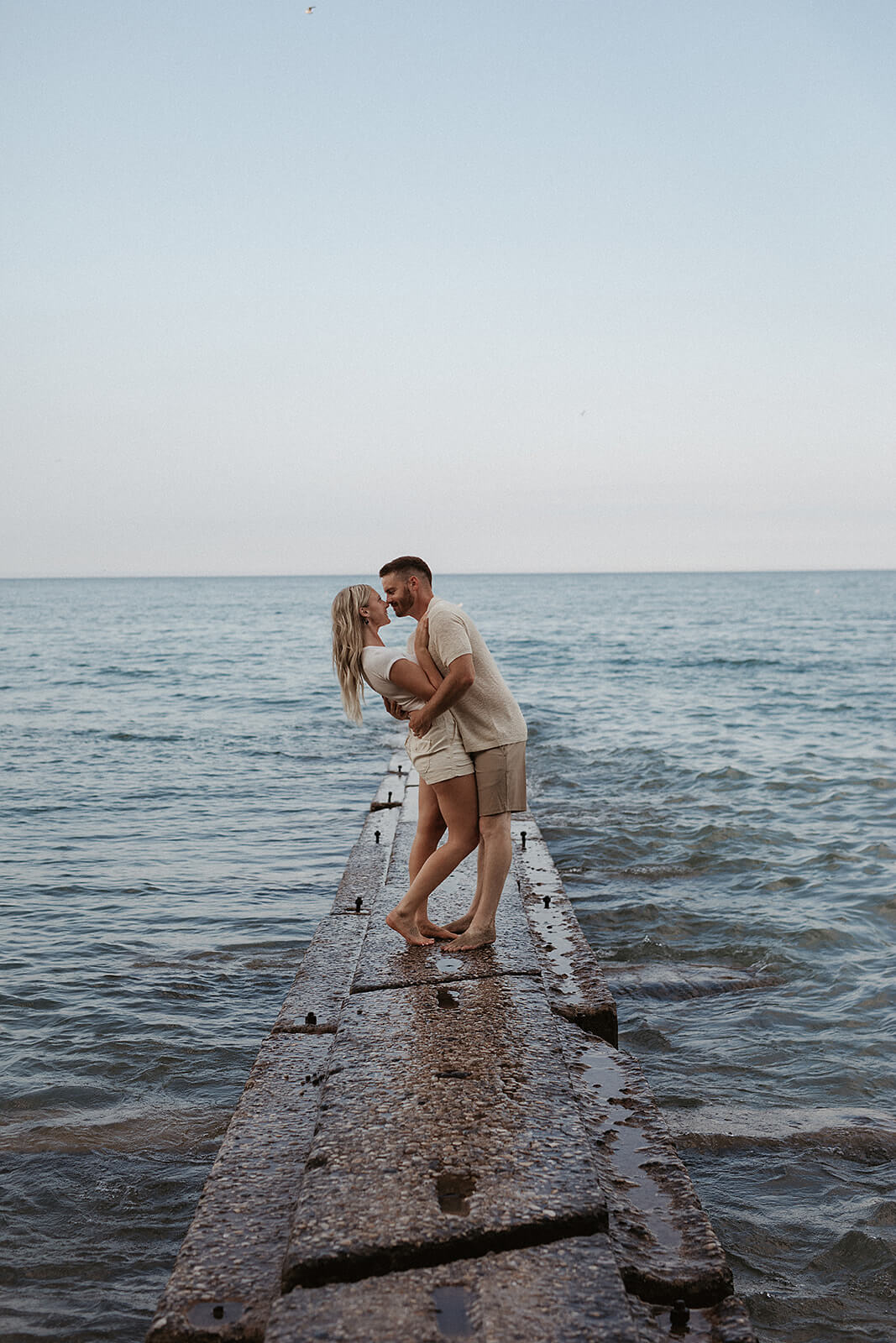 Couple standing together on a concrete pier surrounded by water while embracing near the edge of the lake during sunset.
