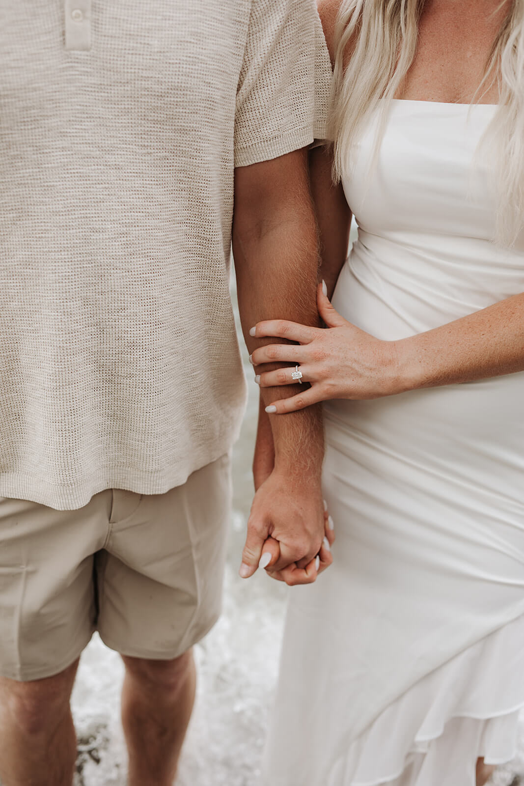 Close-up detail photo of a couple holding hands while standing barefoot near the shoreline, wearing neutral-colored clothing beside the water.