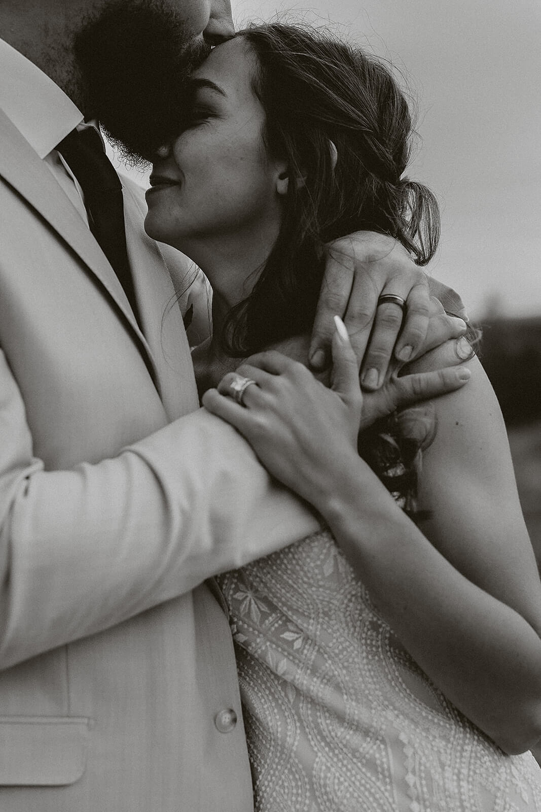 Black and white close-up of a groom kissing his bride on the forehead while she holds his hand showing her wedding ring