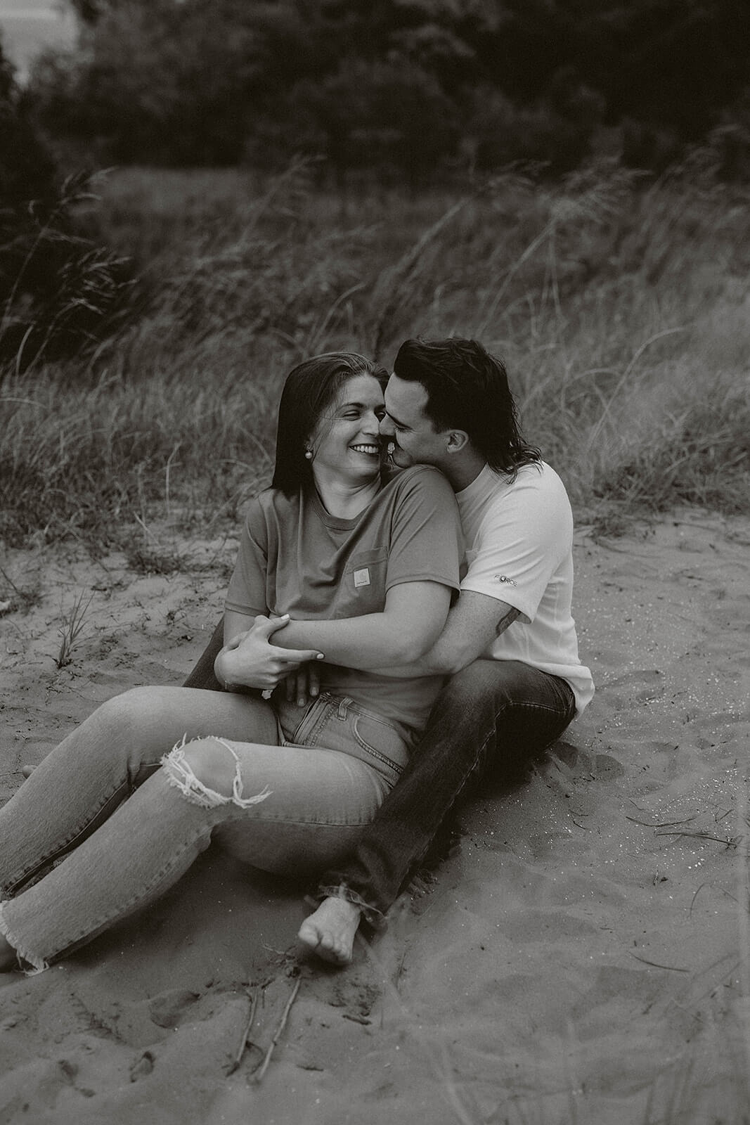 Black and white photo of a couple sitting together on sandy dunes while embracing during an outdoor engagement session.