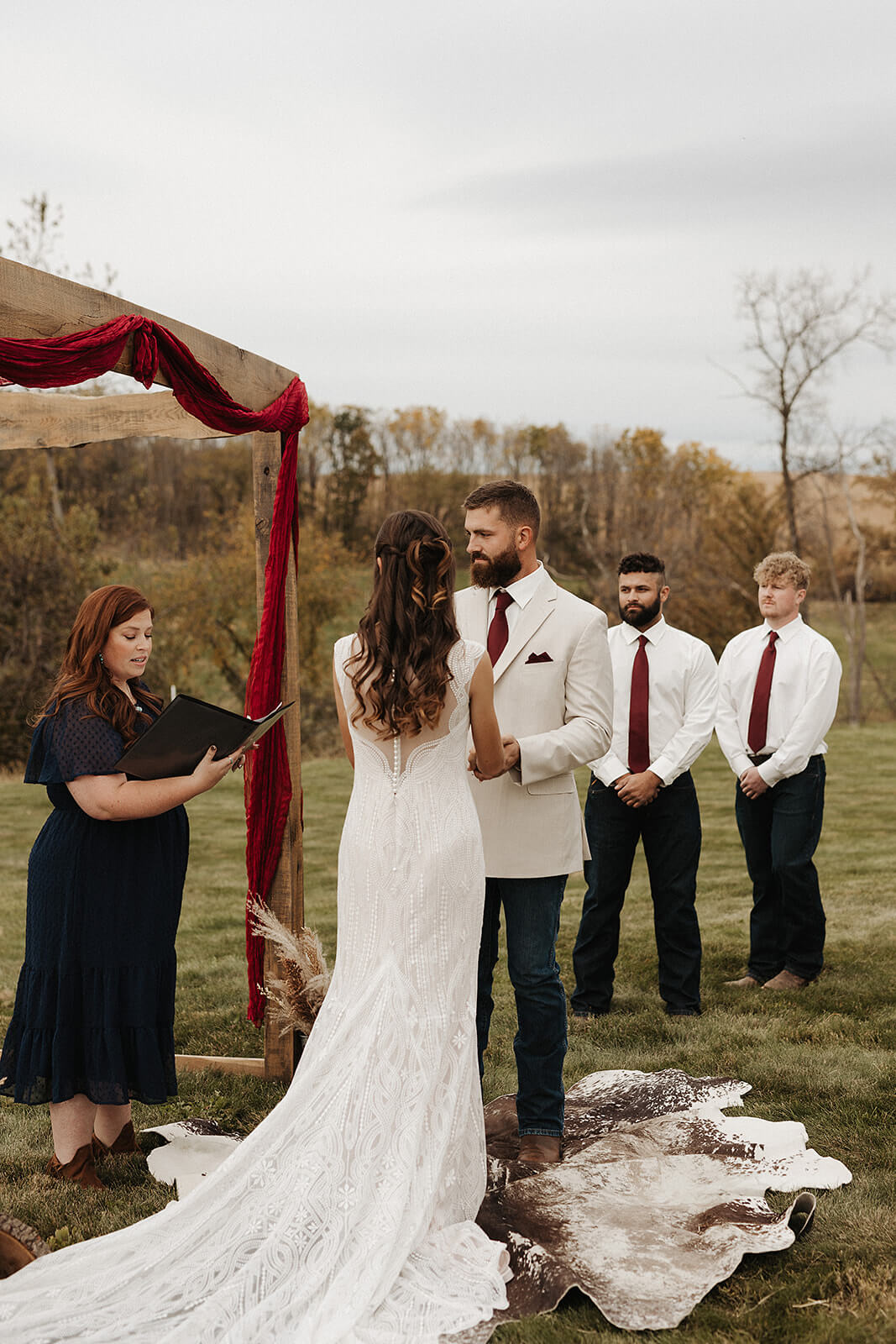 Bride and groom face to face during their outdoor ceremony under a red-draped wooden arch on the rolling hills of a Lancaster Wisconsin family farm