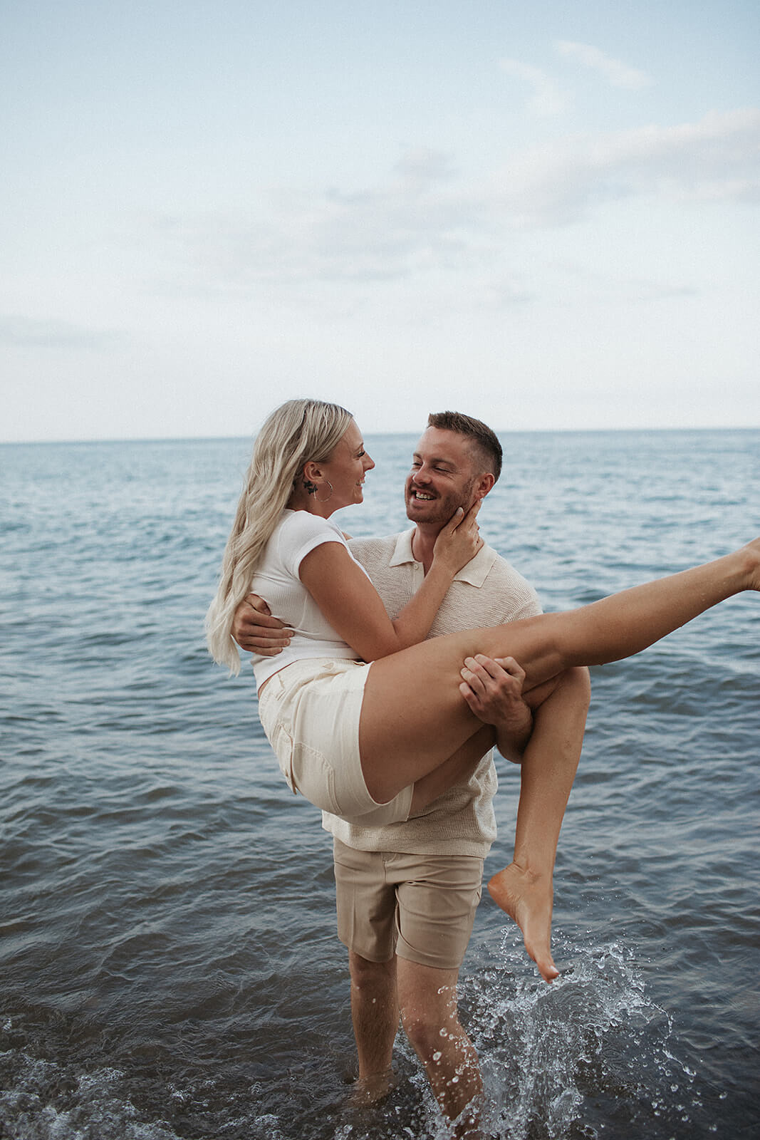 Couple standing together in shallow lake water near the shoreline while laughing and embracing during a beach engagement session.