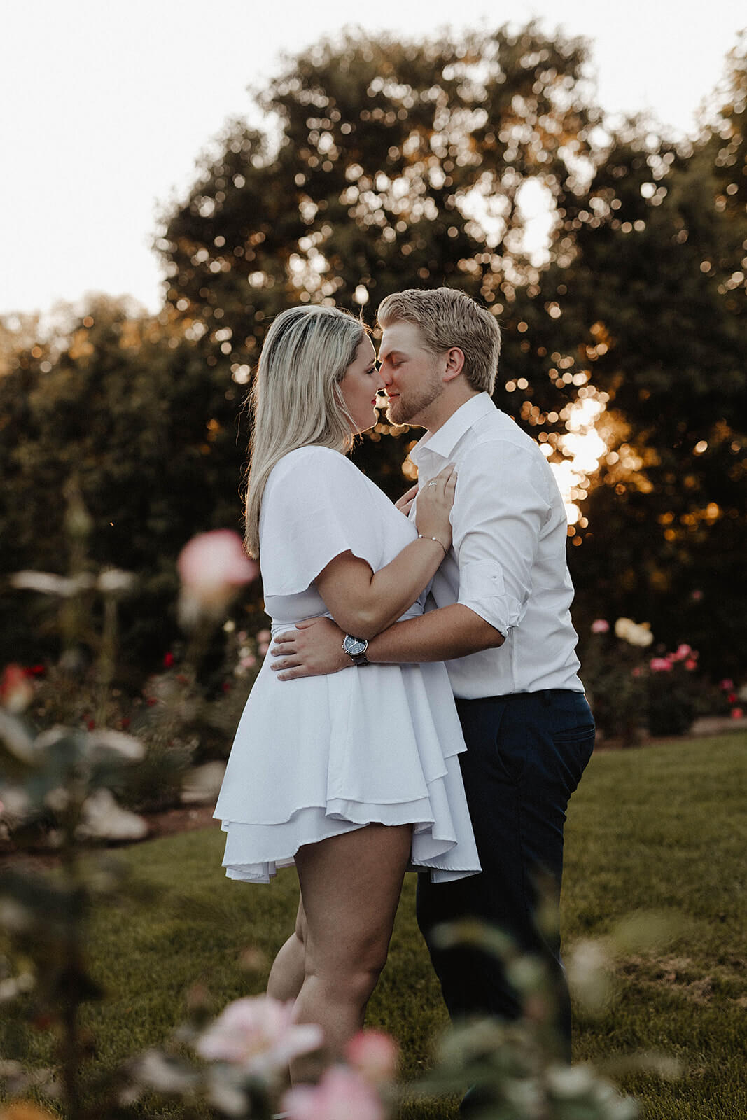 Couple embracing in a grassy garden during sunset with flowers and trees softly blurred in the foreground and background.