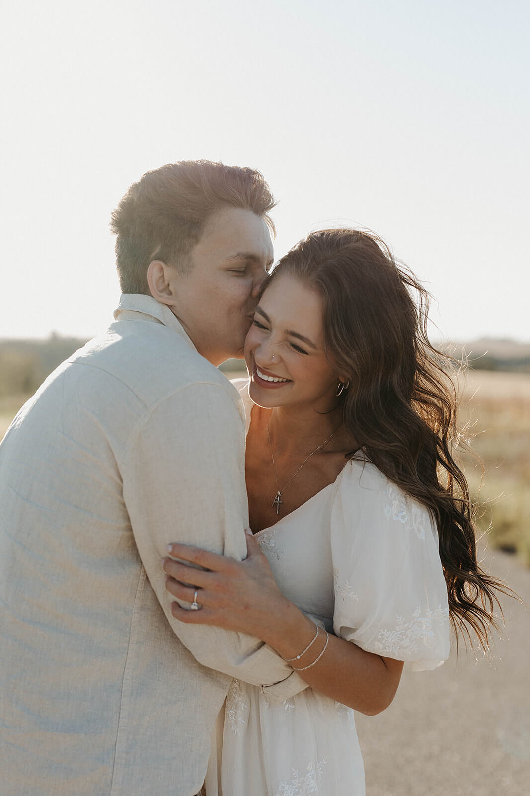 Close-up portrait of a couple embracing and smiling together during golden hour in an open field with soft sunlight surrounding them.