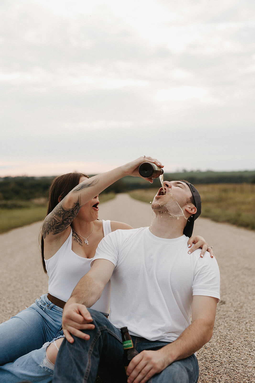 Couple sitting together on a gravel road while laughing and pouring a drink during a playful outdoor engagement session in the countryside.