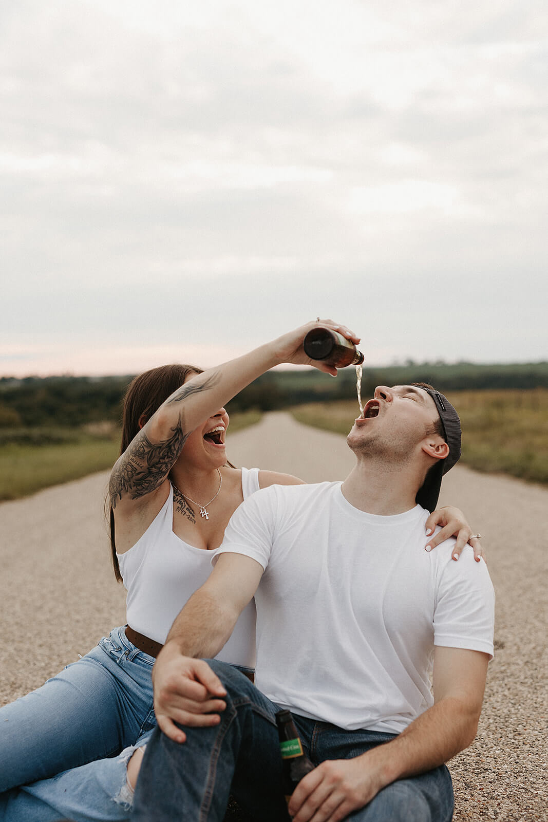 Couple sitting together on a gravel road while laughing and pouring a drink during a playful outdoor engagement session.