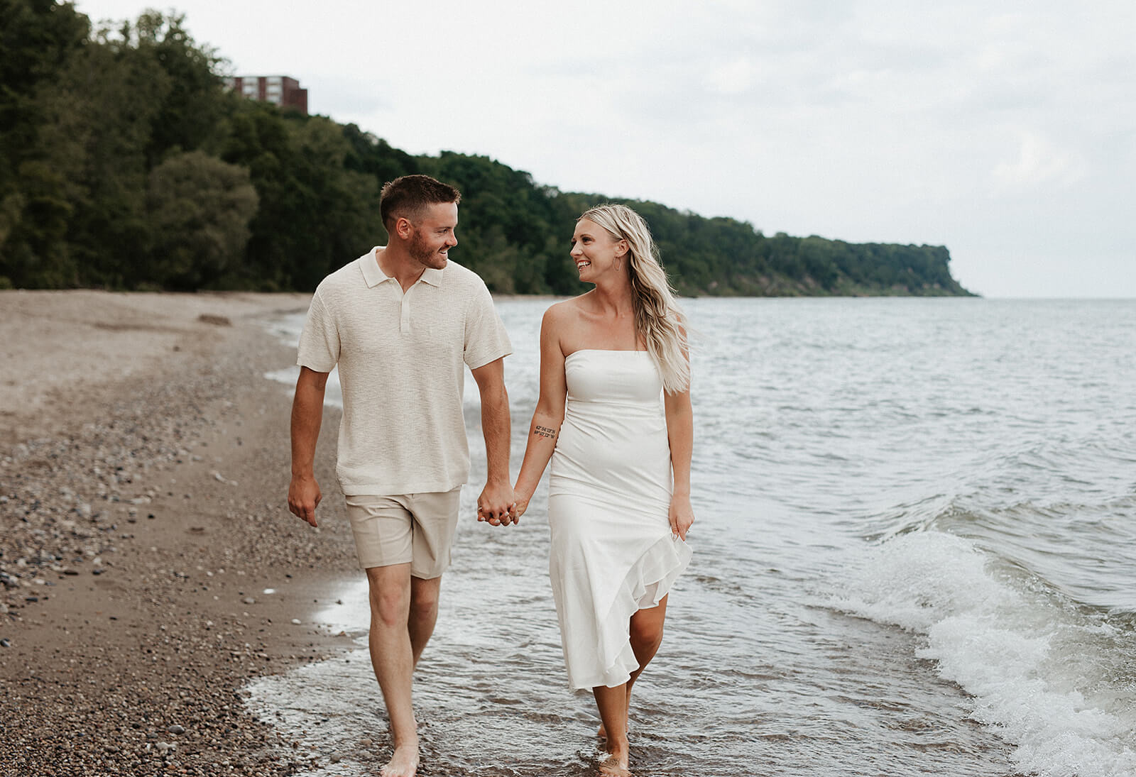 Couple walking hand in hand barefoot along a rocky beach shoreline with small waves washing onto the sand beside them.