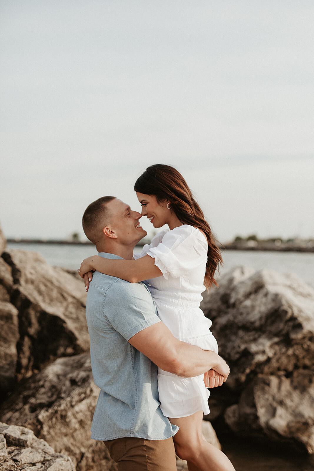 Couple embracing on large rocks near the lake during sunset, with the woman sitting in the man’s arms beside the shoreline.