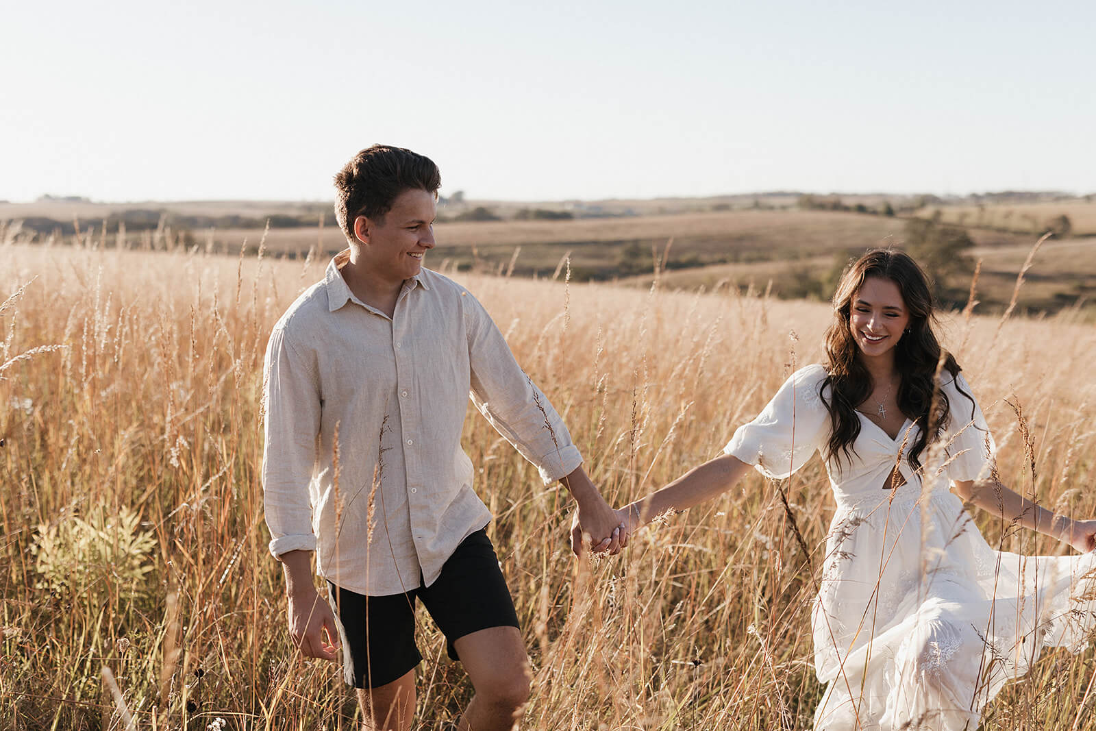 Couple walking hand in hand through a tall golden grass field during sunset, wearing light neutral clothing with rolling hills in the background.