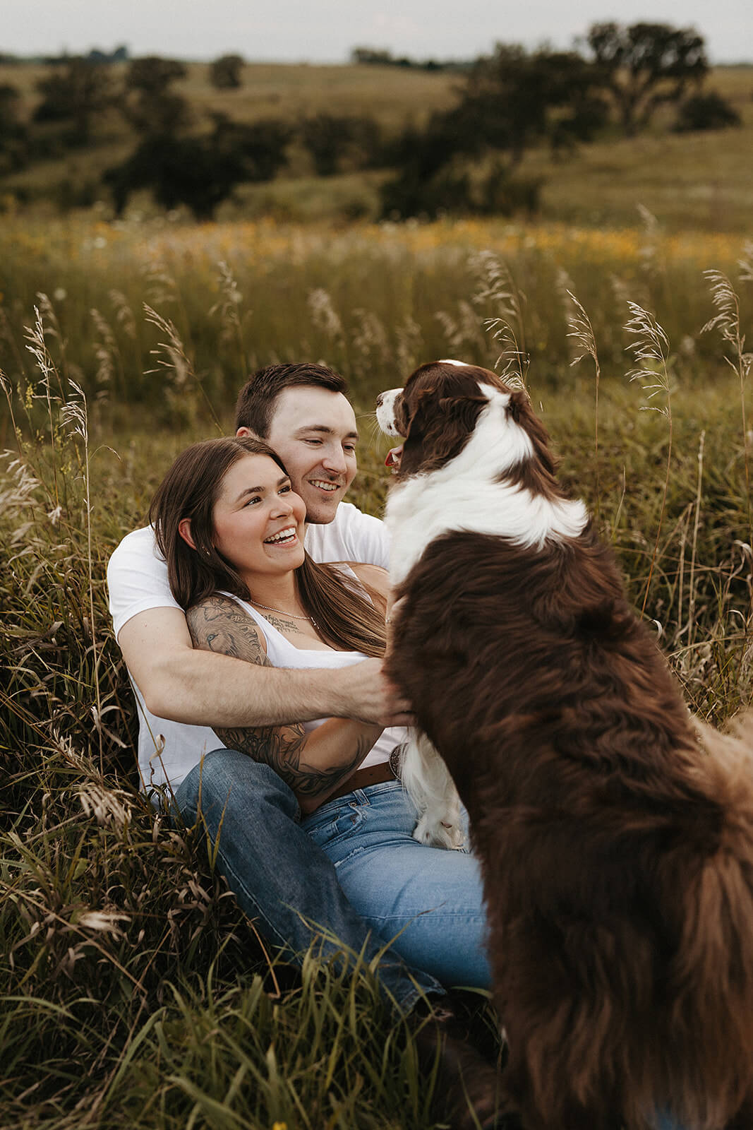 Couple sitting together in a grassy field with their dog standing in front of them during golden hour, with tall grass and open countryside surrounding them.
