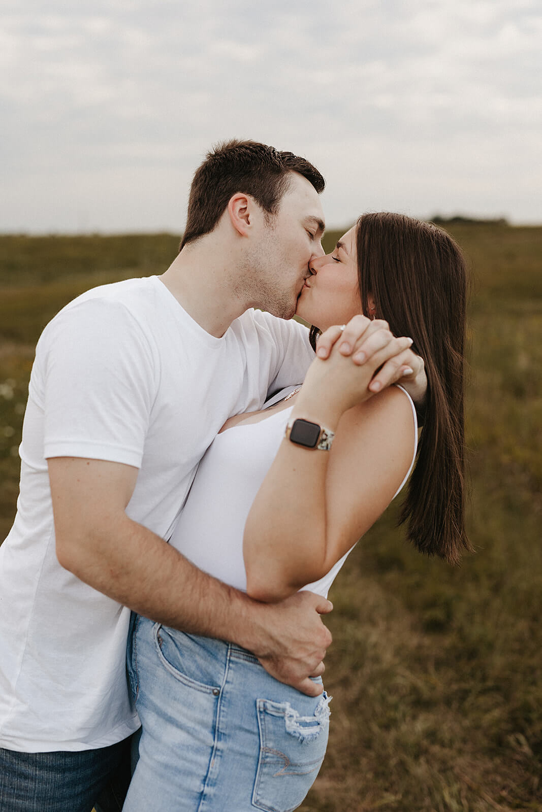Couple embracing and kissing in an open grassy field during golden hour, wearing casual white clothing with rolling hills in the background.
