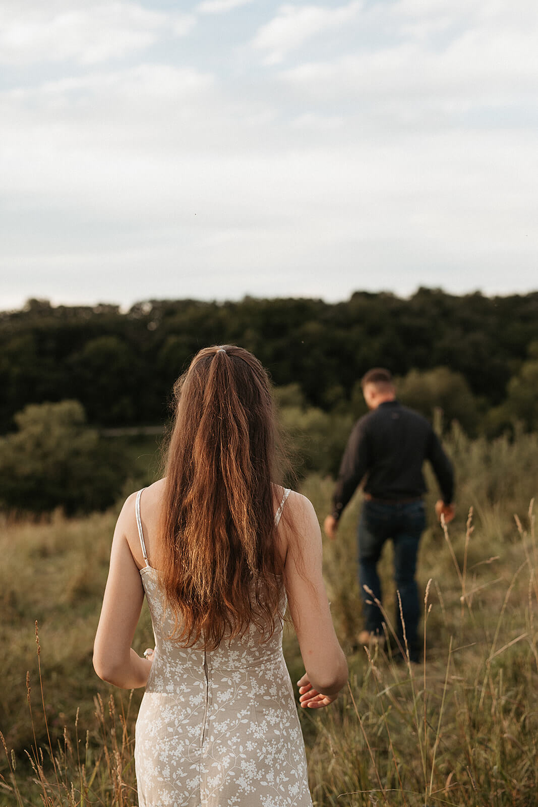 Woman walking through a grassy field toward her partner during sunset with rolling hills and trees in the background.