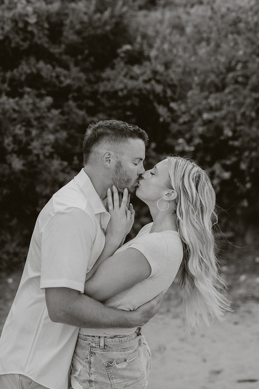 Black and white portrait of a couple embracing and kissing outdoors near the beach with trees blurred in the background.