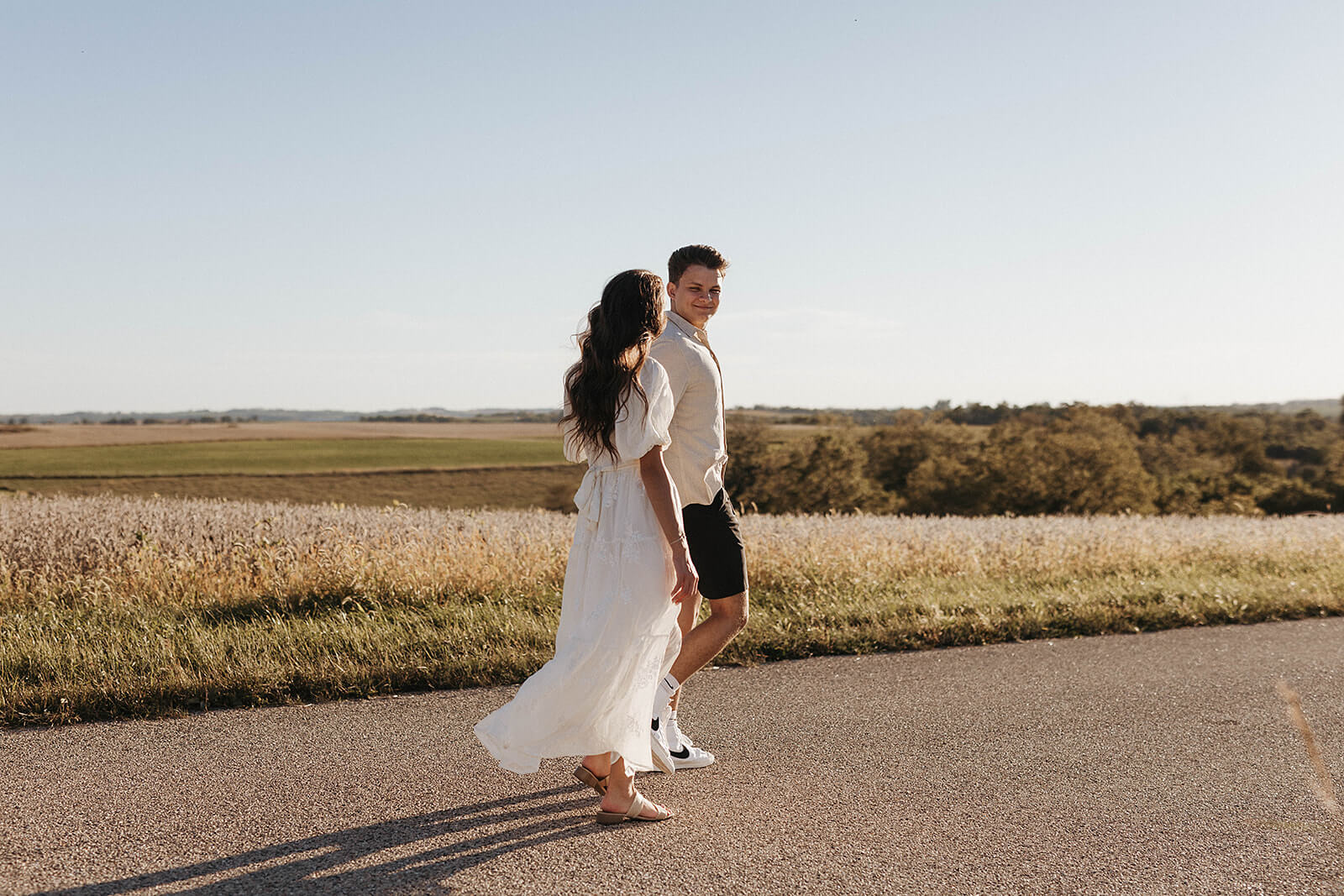 Couple walking together on a quiet country road beside open grassy fields during golden hour, wearing light neutral clothing.