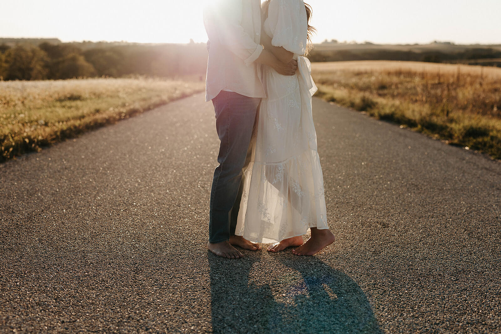 Couple embracing barefoot in the middle of a country road during golden hour with long shadows cast across the pavement and sunlight behind them.