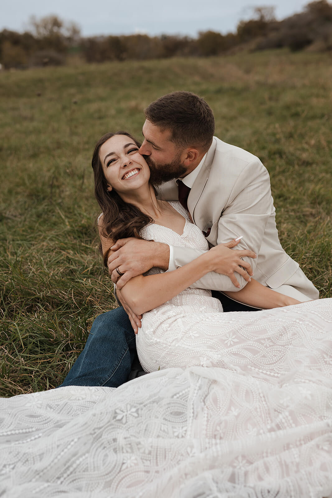 Bride and groom sitting together in a green farm field laughing and embracing during Lancaster Wisconsin wedding portraits