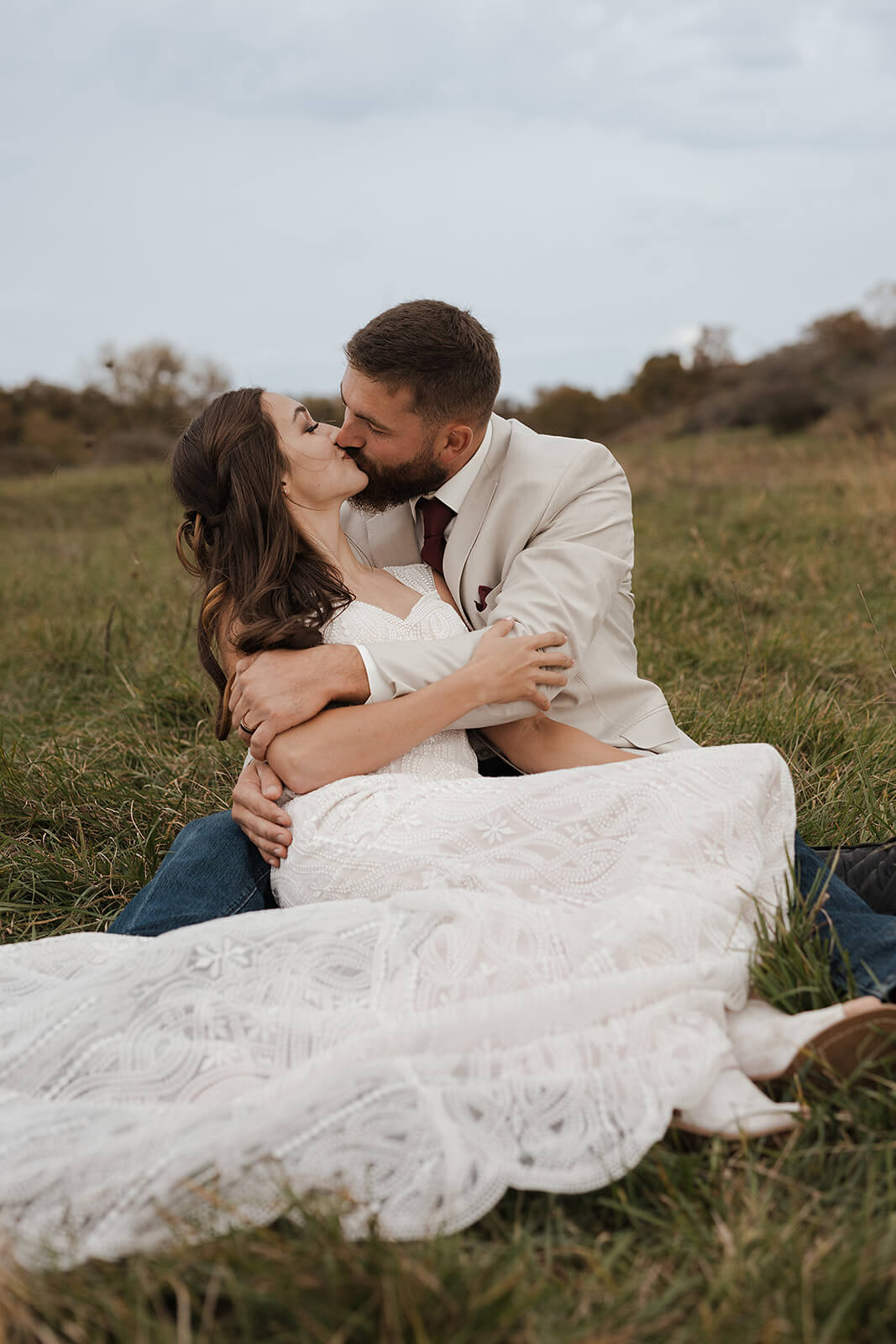 Bride and groom sitting together in a green farm field laughing and embracing during Lancaster Wisconsin wedding portraits