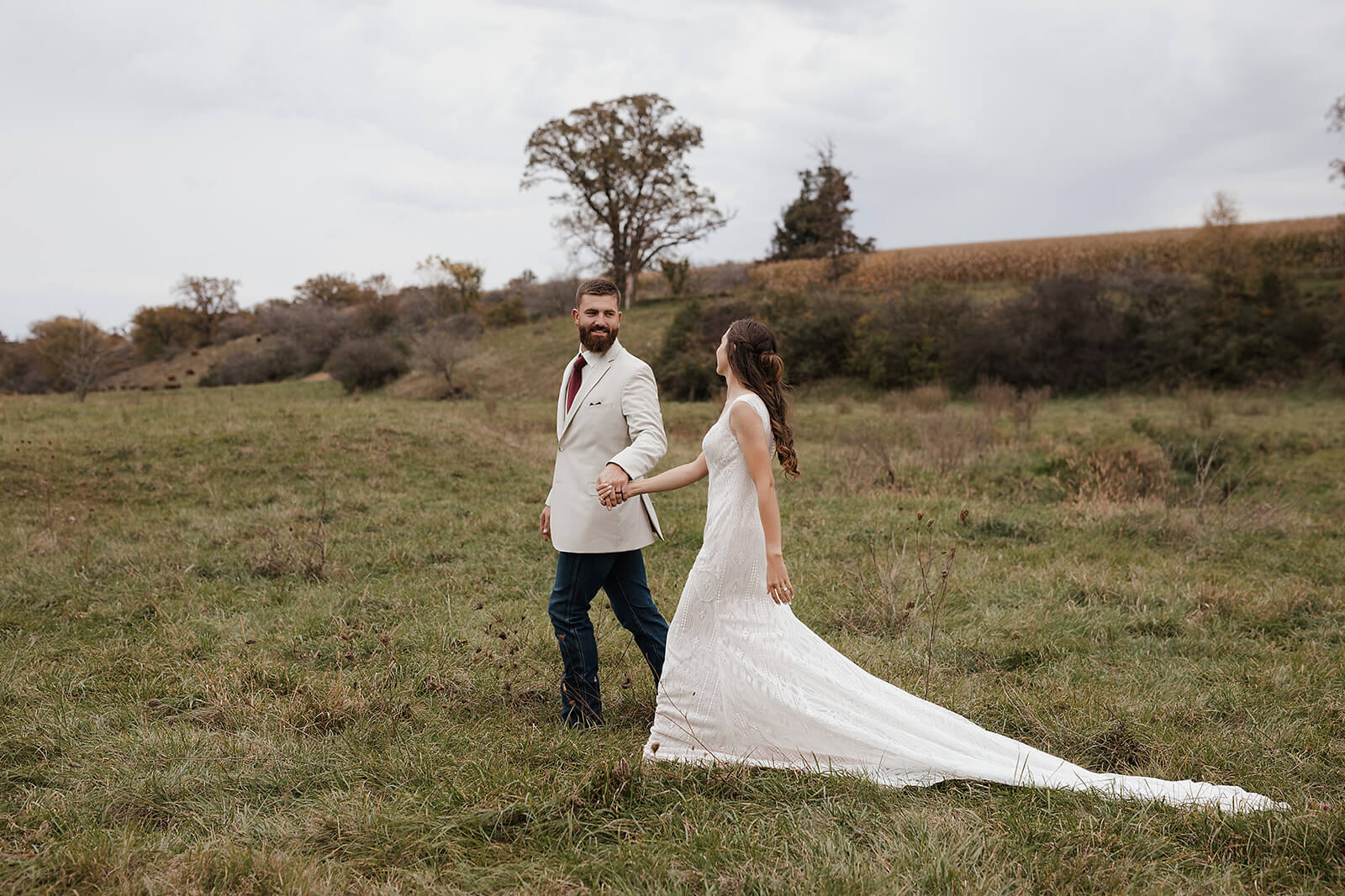Groom leading his bride by the hand through a wide open green field with rolling hills in the background during Lancaster Wisconsin wedding portraits
