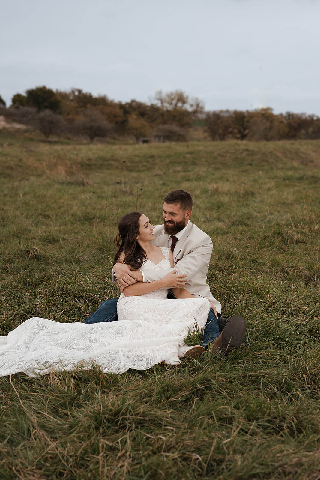 Bride and groom sitting together in a green farm field laughing during Lancaster Wisconsin wedding portraits