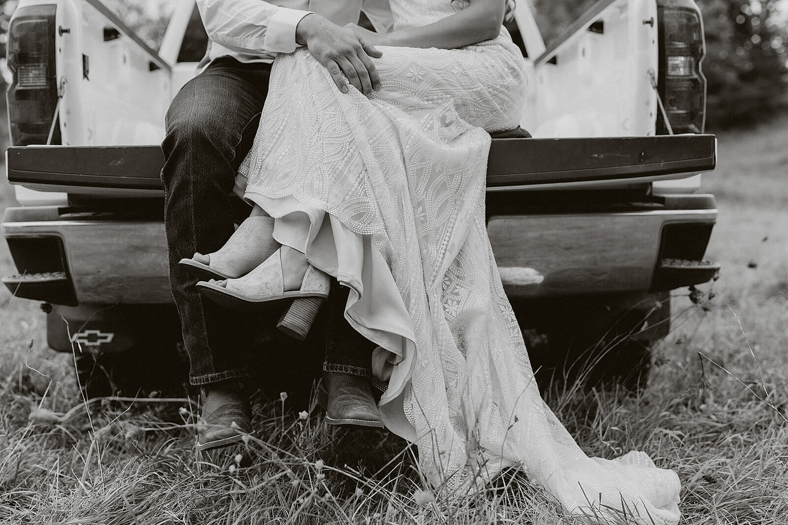 Black and white close-up of a bride in a lace gown and cowboy boots sitting with her groom in the back of a pickup truck at a Lancaster Wisconsin farmhouse wedding
