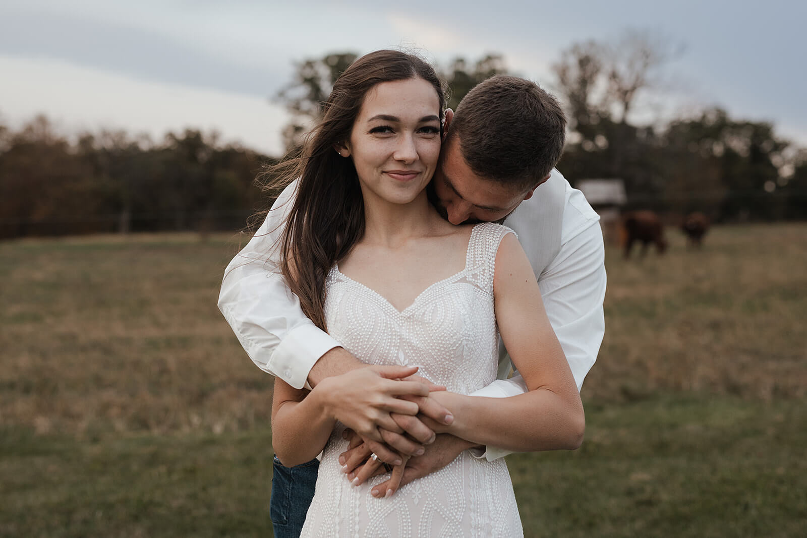 Bride smiling at the camera while the groom kisses her cheek during farm wedding portraits with cattle visible in the background at a Lancaster Wisconsin wedding