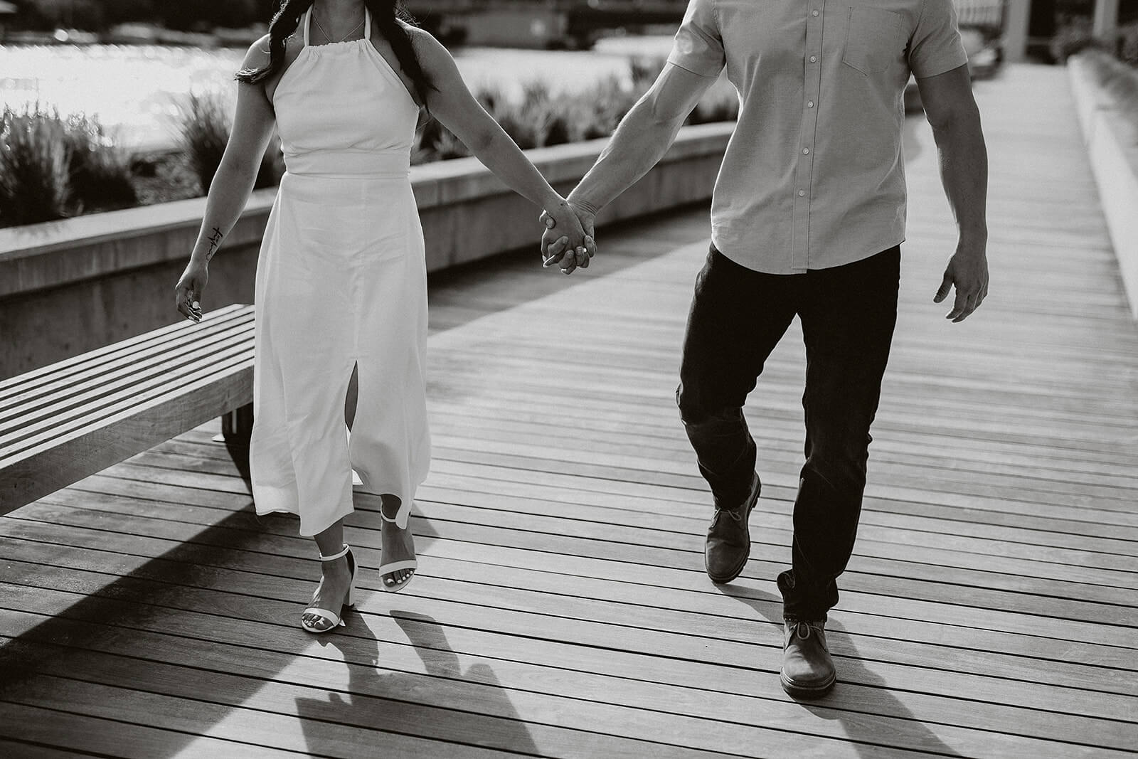 Black and white photo of a couple holding hands while walking together on a wooden boardwalk near the waterfront.