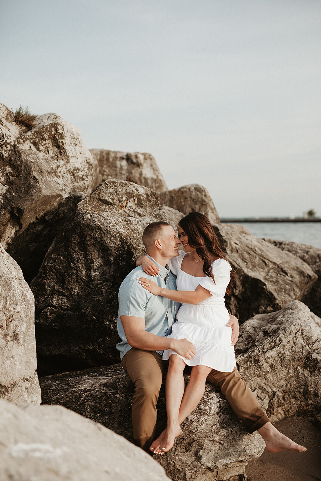 Couple sitting together on large shoreline rocks while embracing near the water during a beach engagement session.