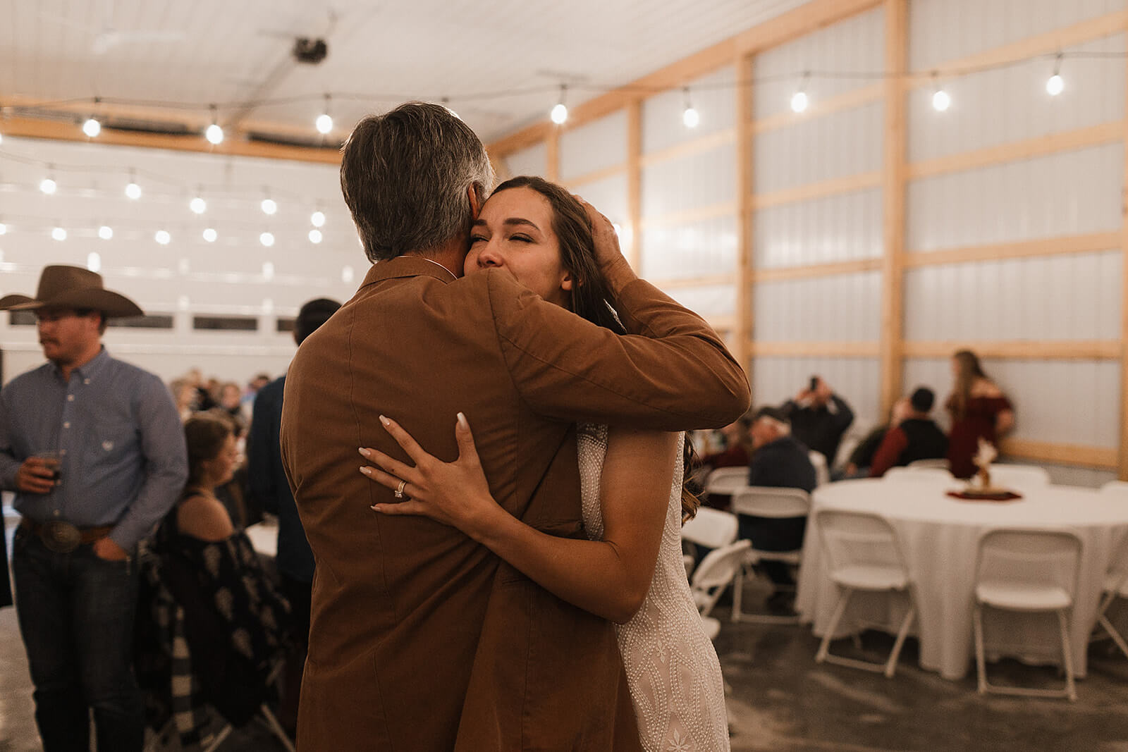Bride hugging her father during the reception inside a Lancaster Wisconsin barn wedding venue with string lights overhead