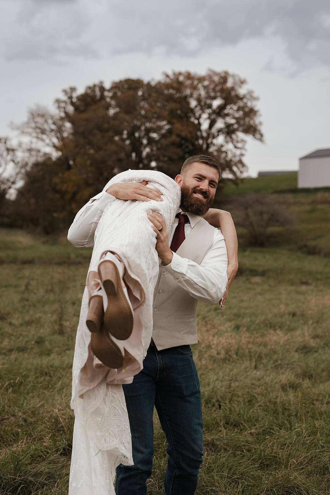 Groom laughing while carrying his bride over his shoulder in a farm field during Lancaster Wisconsin wedding portraits