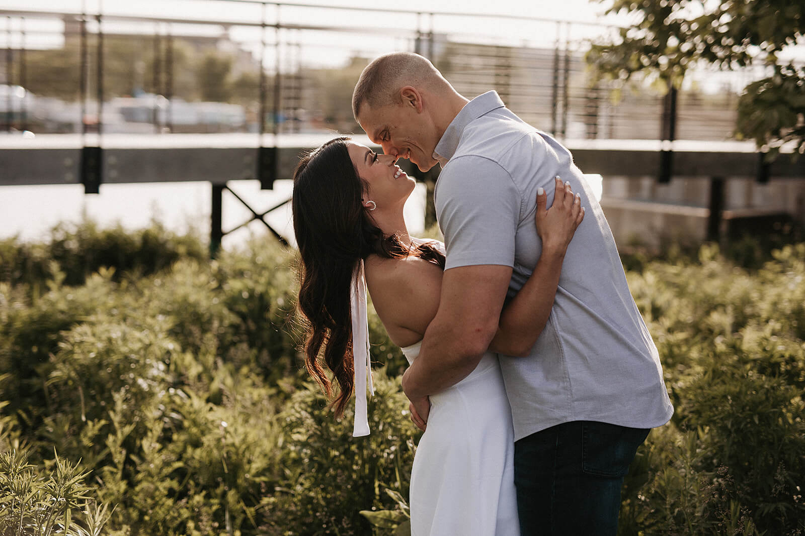 Couple embracing outdoors near a waterfront garden with greenery and a bridge structure blurred in the background.